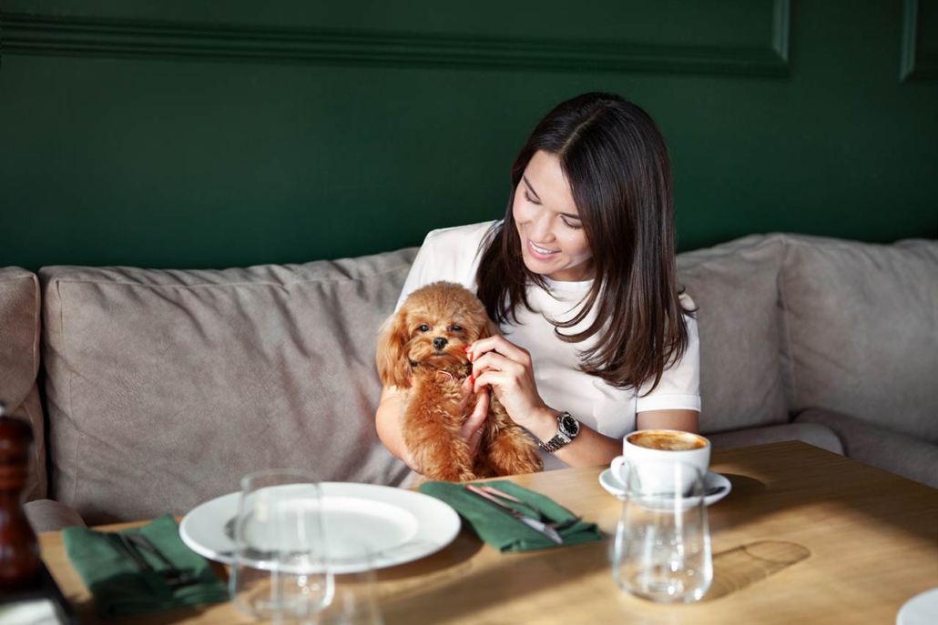 Beautiful smiling Woman holds small dog in hands sits at table in restaurant with cup of coffee