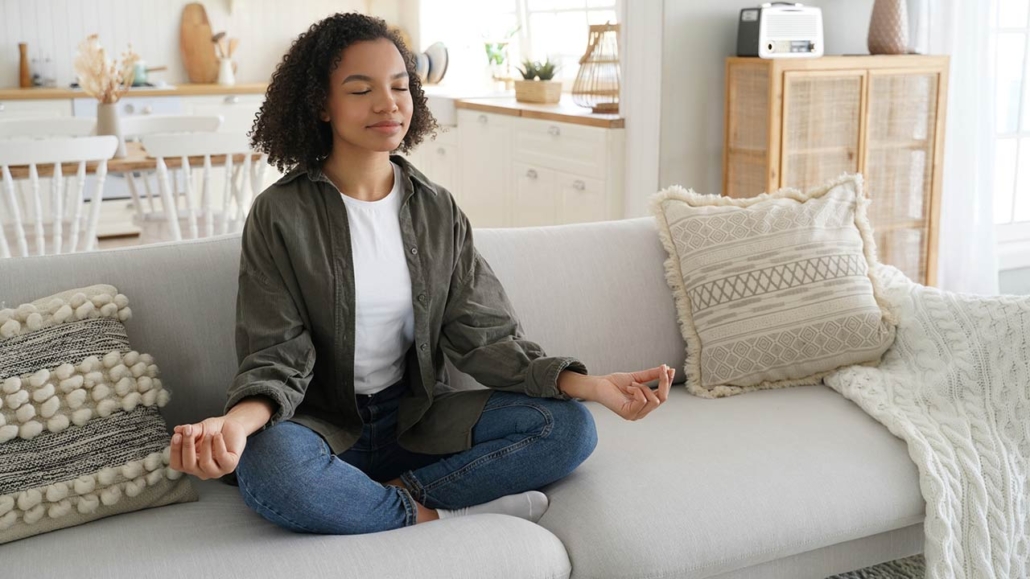 A serene Black woman meditates on a couch, eyes closed, in a sunlit, stylish living room