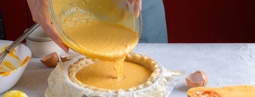 Woman add a pumpkin filling puree into a baking dish for cooking pumpkin pie on a gray table with ingredients in the kitchen