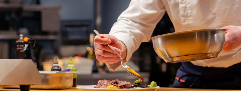 close up chef in the kitchen garnishes medium rare sliced beef steak with sauce on a white plate with a spoon