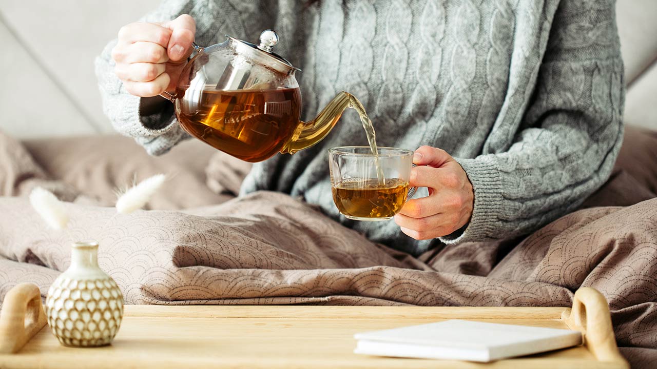 Woman dressed in knitted sweater pours hot tea from glass teapot into mug while sitting in bed