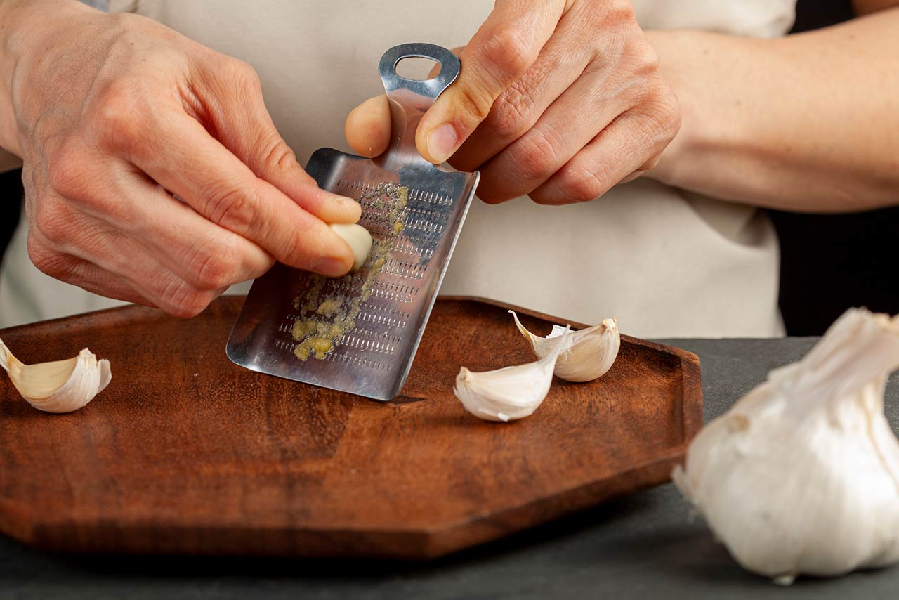 Hands of a caucasian woman grating garlic cloves using shovel shaped metal mini grater on a wooden plate